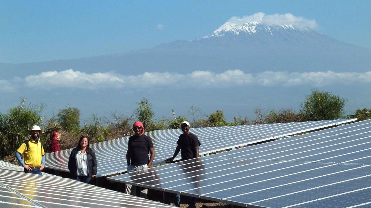 一群太阳能工程师站在山脚下的一块太阳能电池板地里. 乞力马扎罗山是利乐全球最大体育平台主导的电力非洲项目的一部分